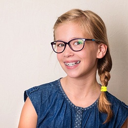 A young girl wearing a denim blouse and glasses showing off her smile that is donning metal braces